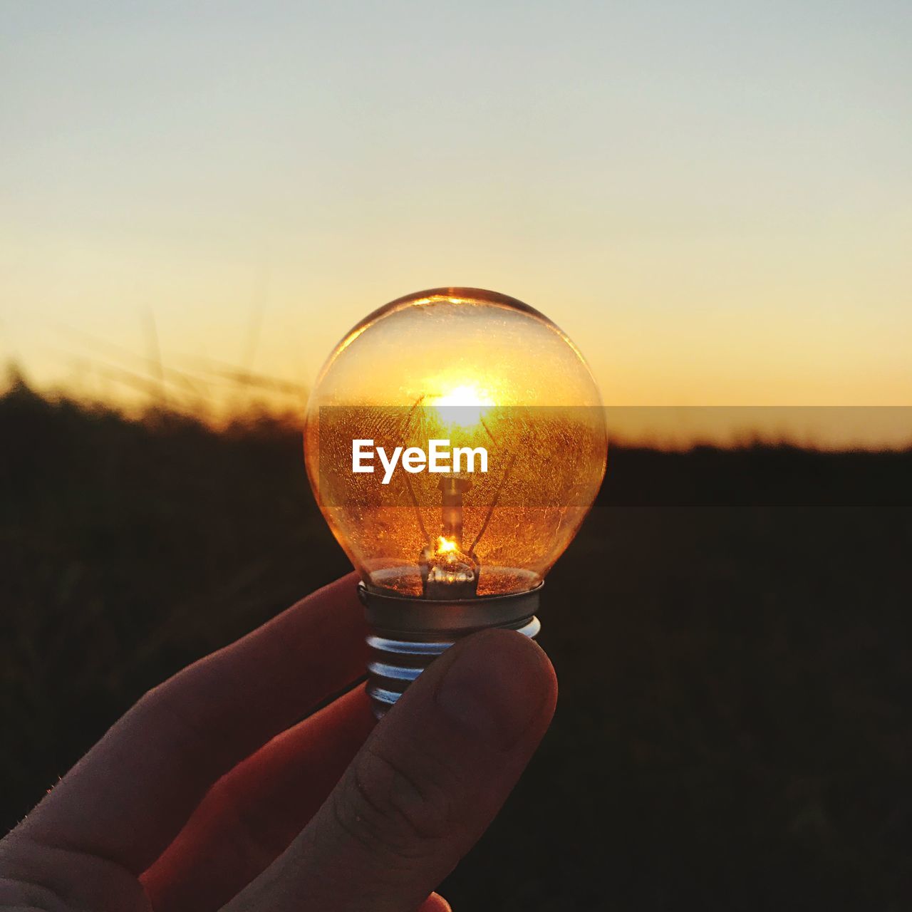 Close-up of hand holding light bulb against sky during sunset