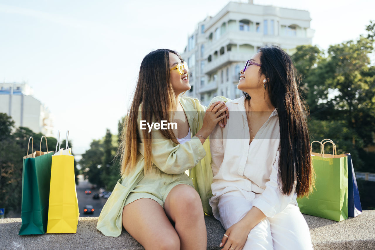 Smiling friends looking at each other while sitting on retaining wall