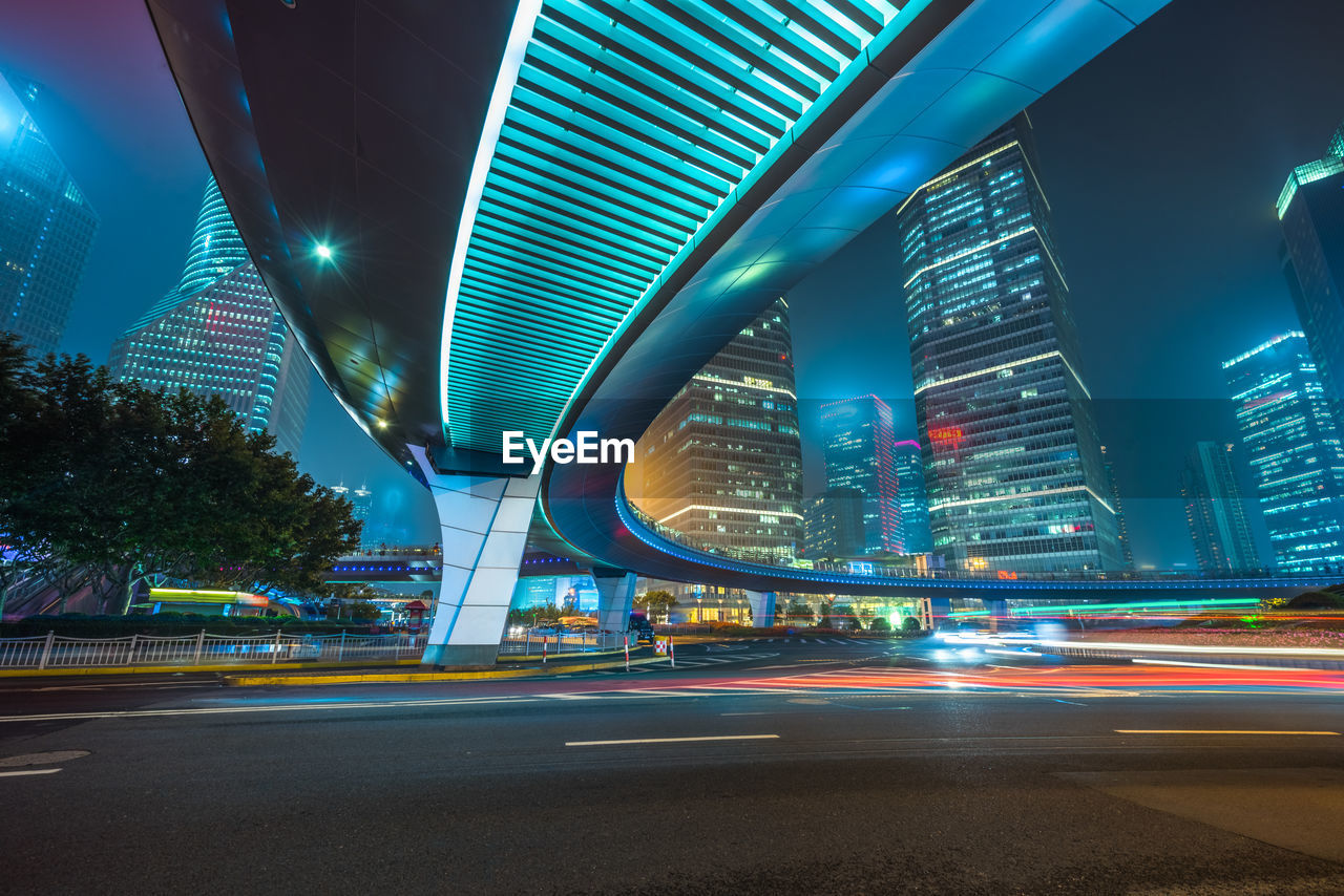 LIGHT TRAILS ON ROAD AMIDST ILLUMINATED BUILDINGS AT NIGHT