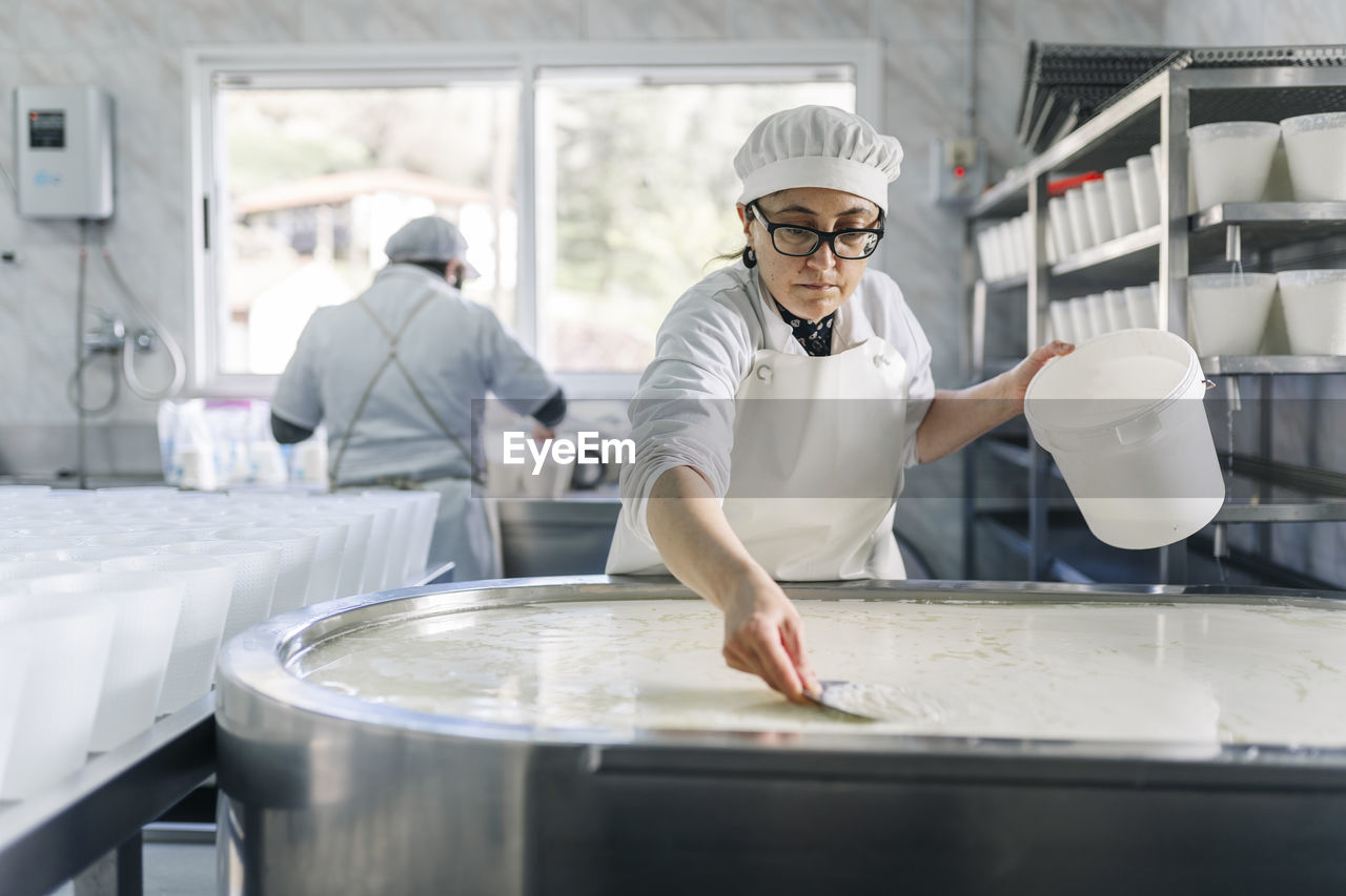 Female chef making cheese with colleague in background at factory