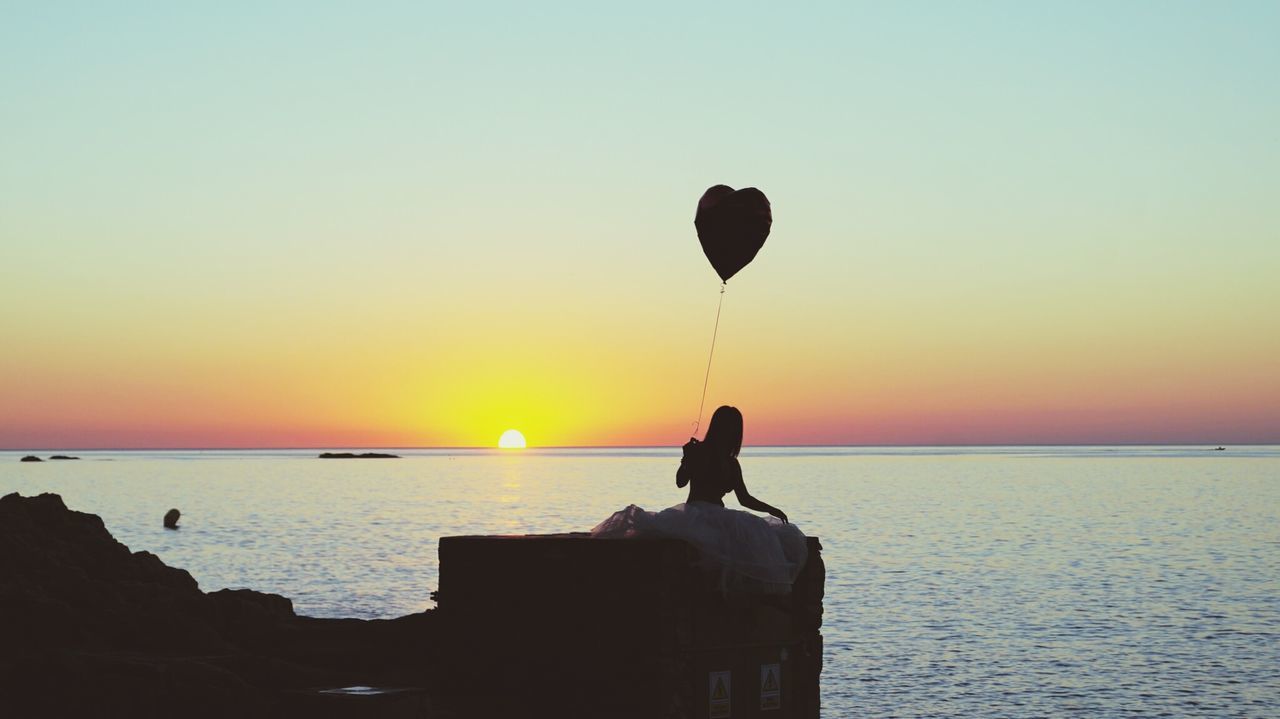 Woman holding heart shape balloon while sitting on retaining wall at sea against sky during sunset