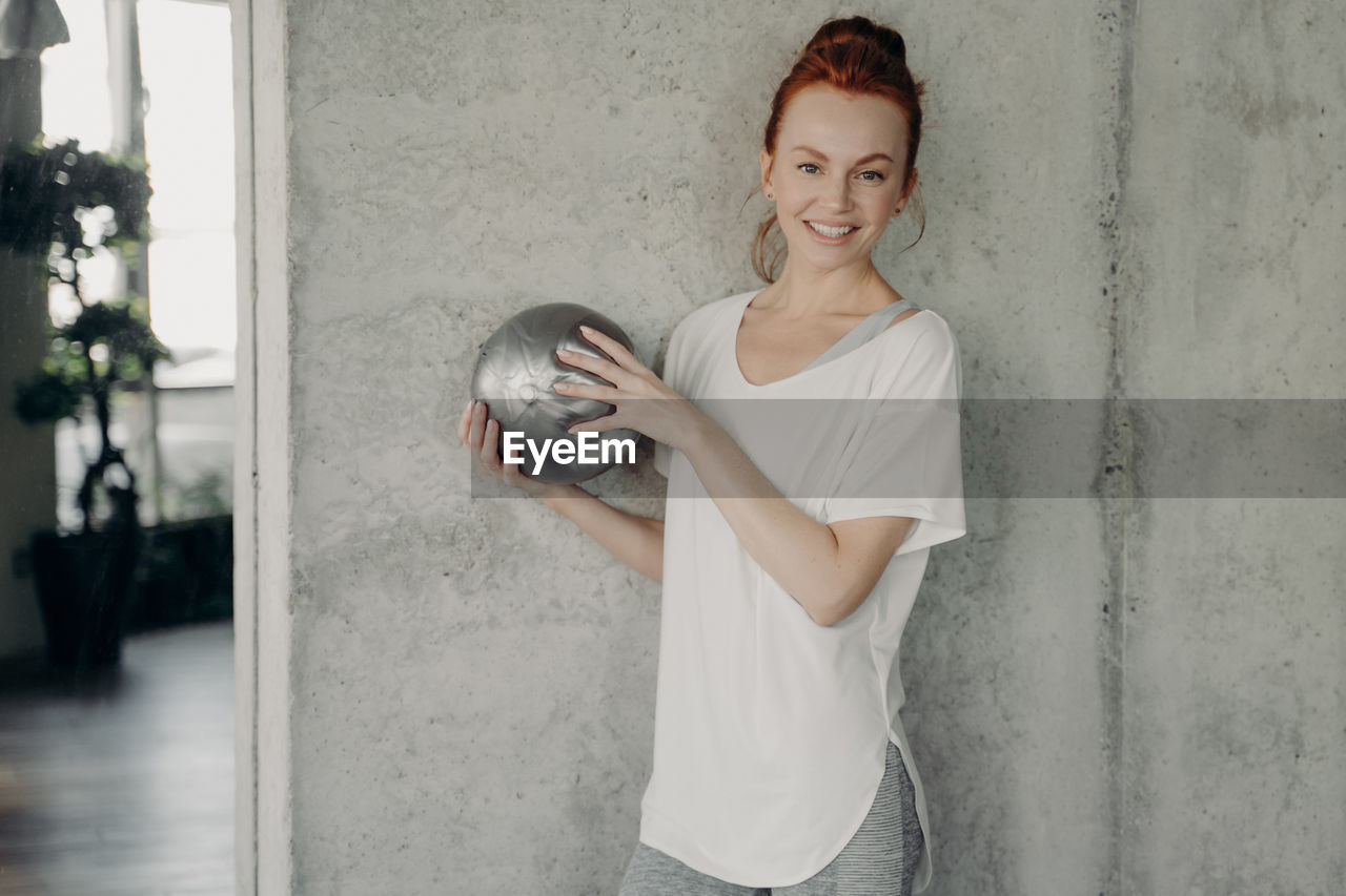 PORTRAIT OF A SMILING YOUNG WOMAN STANDING BY WALL