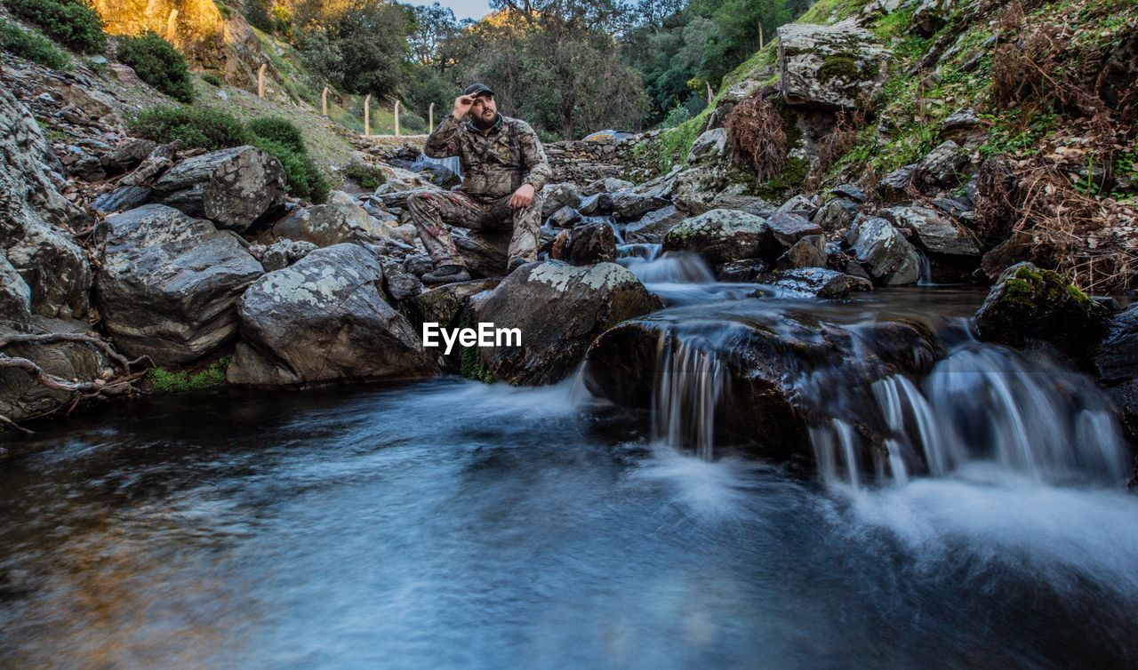 Full length of thoughtful man sitting on rock by stream in forest