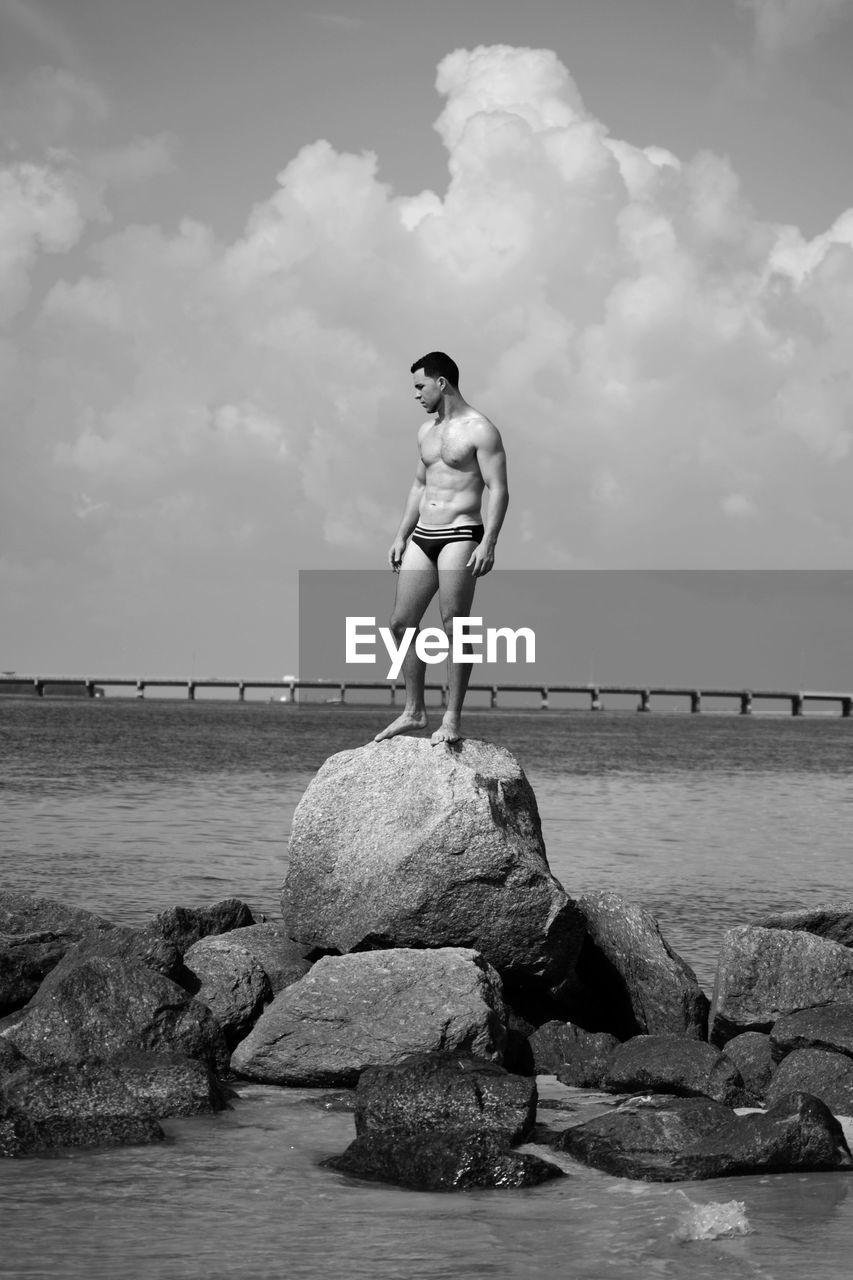 Full length of shirtless man in underwear standing on rock amidst sea against sky
