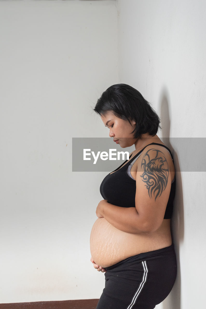 Pregnant woman with hands on stomach standing against wall at home