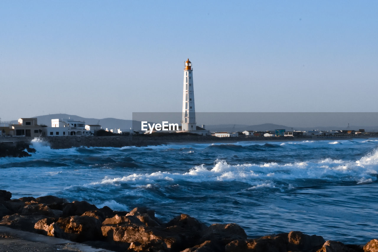 LIGHTHOUSE BY SEA AGAINST BUILDINGS AGAINST CLEAR SKY