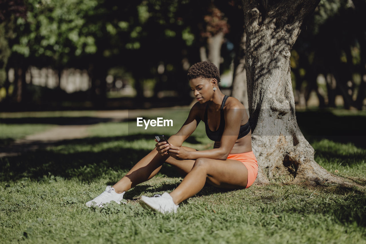 African american woman in sportswear holding smartphone while sitting on grass in sunny day