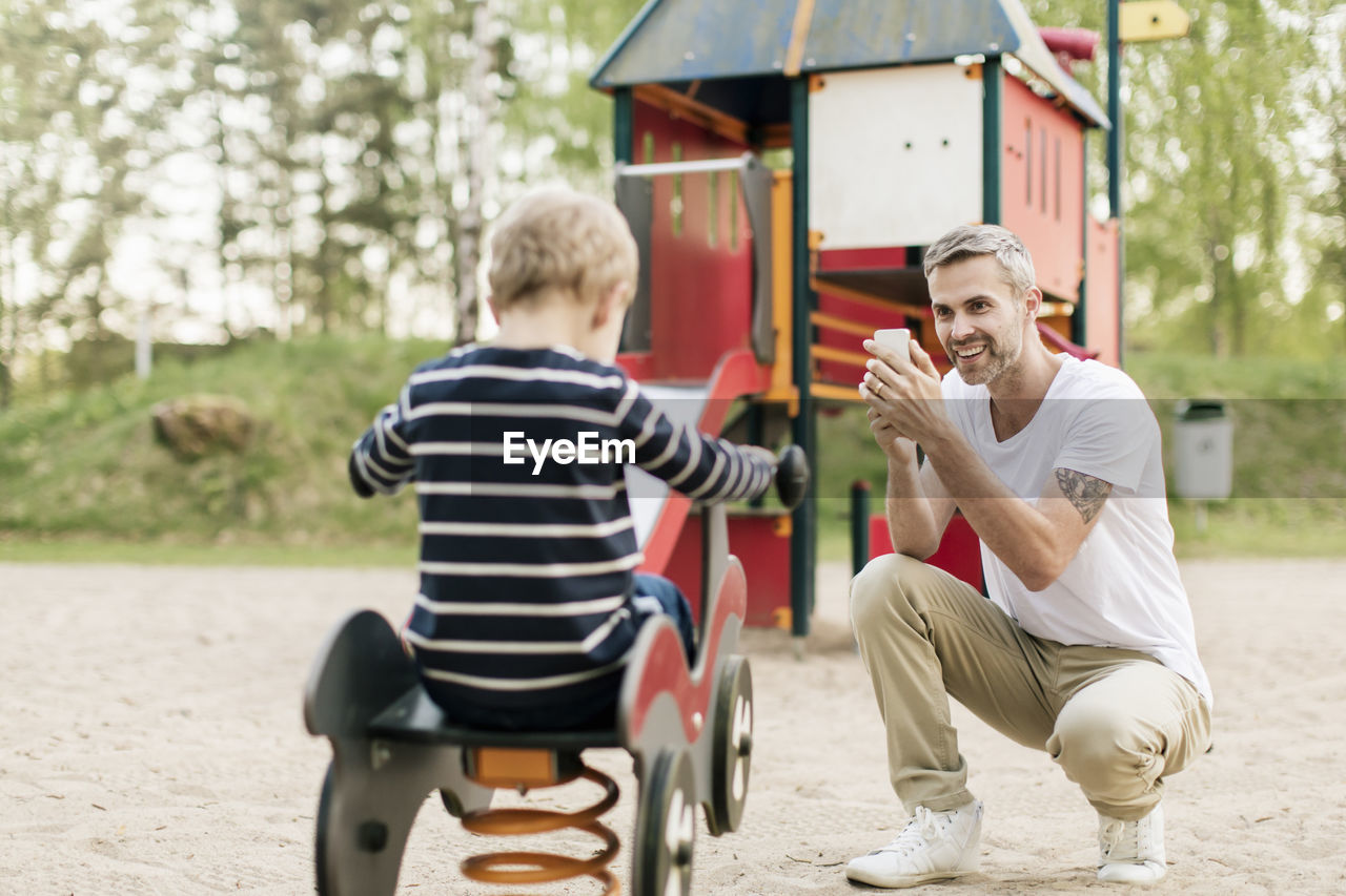 Smiling father photographing son playing on spring ride at playground