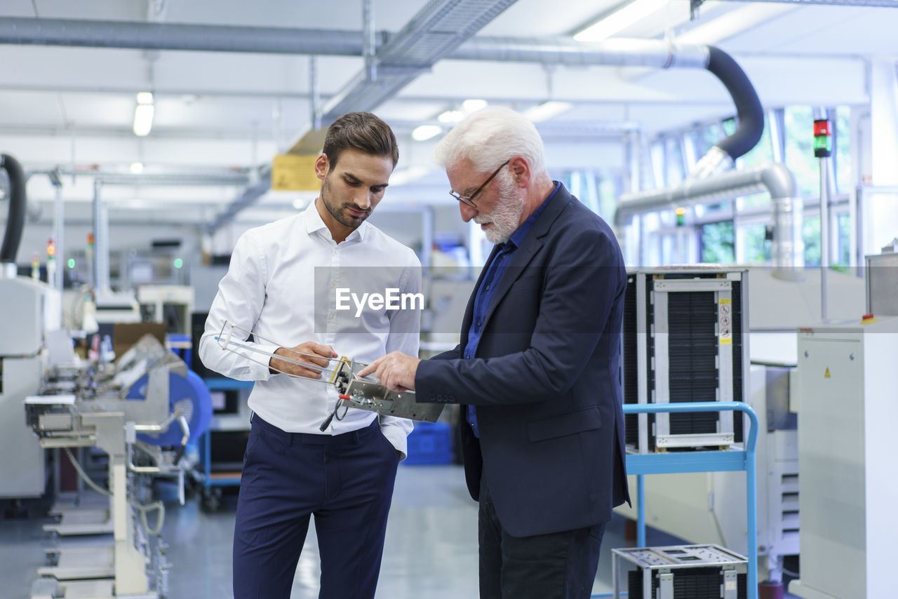 Senior manager pointing at machine part while discussing with engineer by machinery in factory