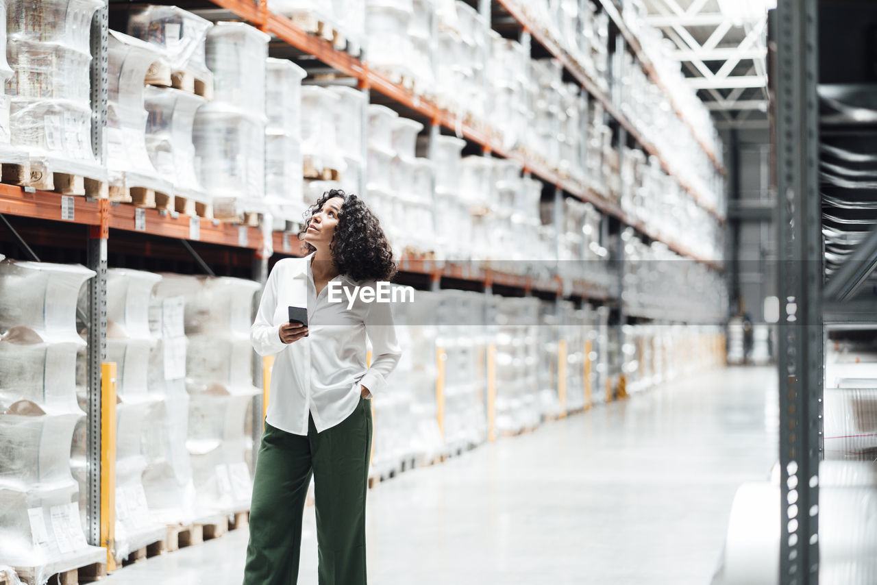 Businesswoman with smart phone standing by rack in warehouse