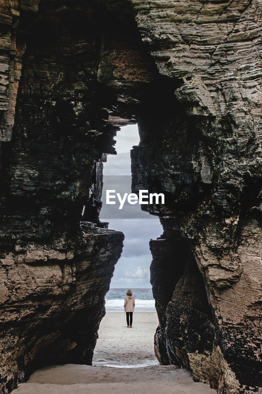 View of distant anonymous traveler in outerwear through massive stony formation on sandy coast near sea in nature of spain