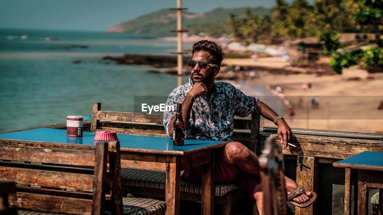 Young man wearing sunglasses while sitting on bench by sea during summer