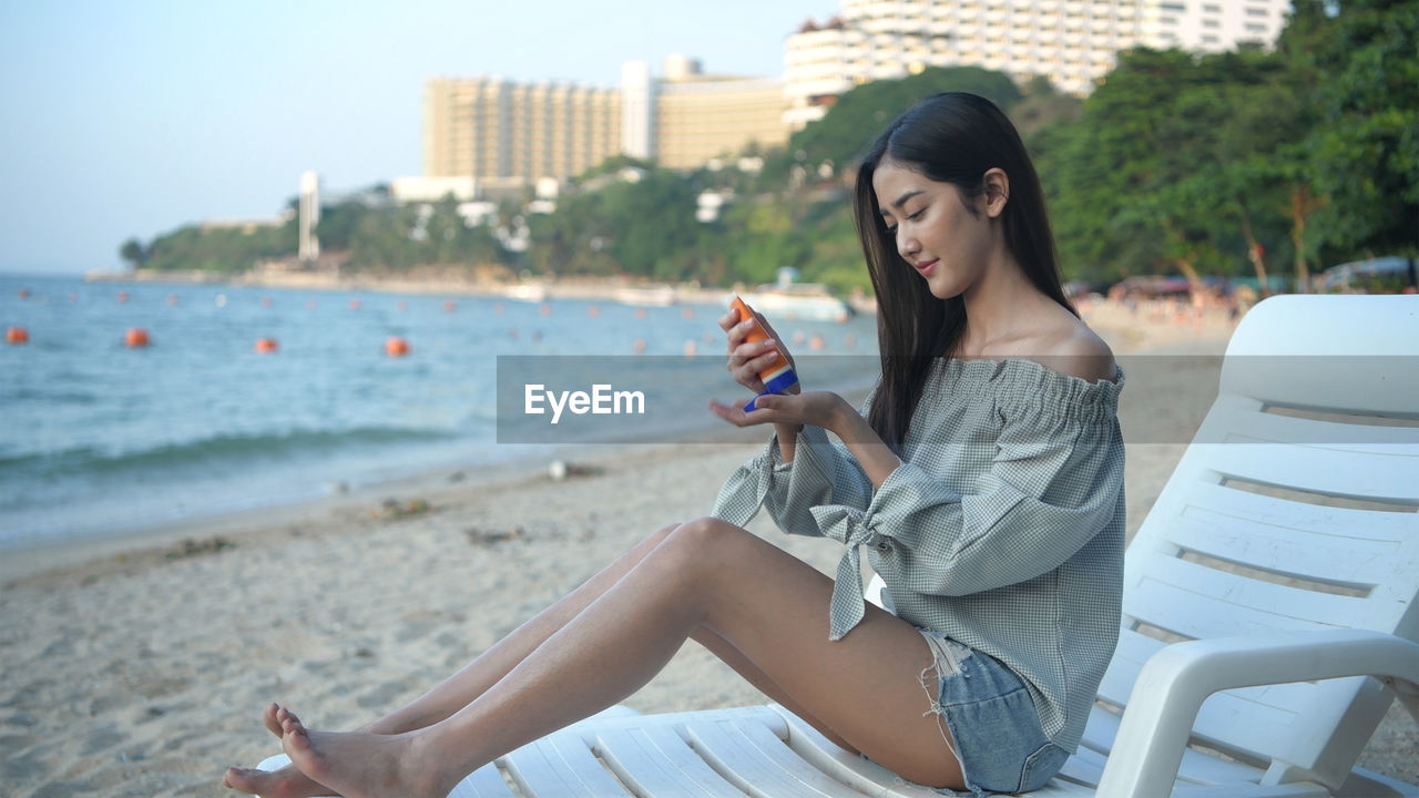 YOUNG WOMAN USING MOBILE PHONE WHILE SITTING ON LAND AT BEACH