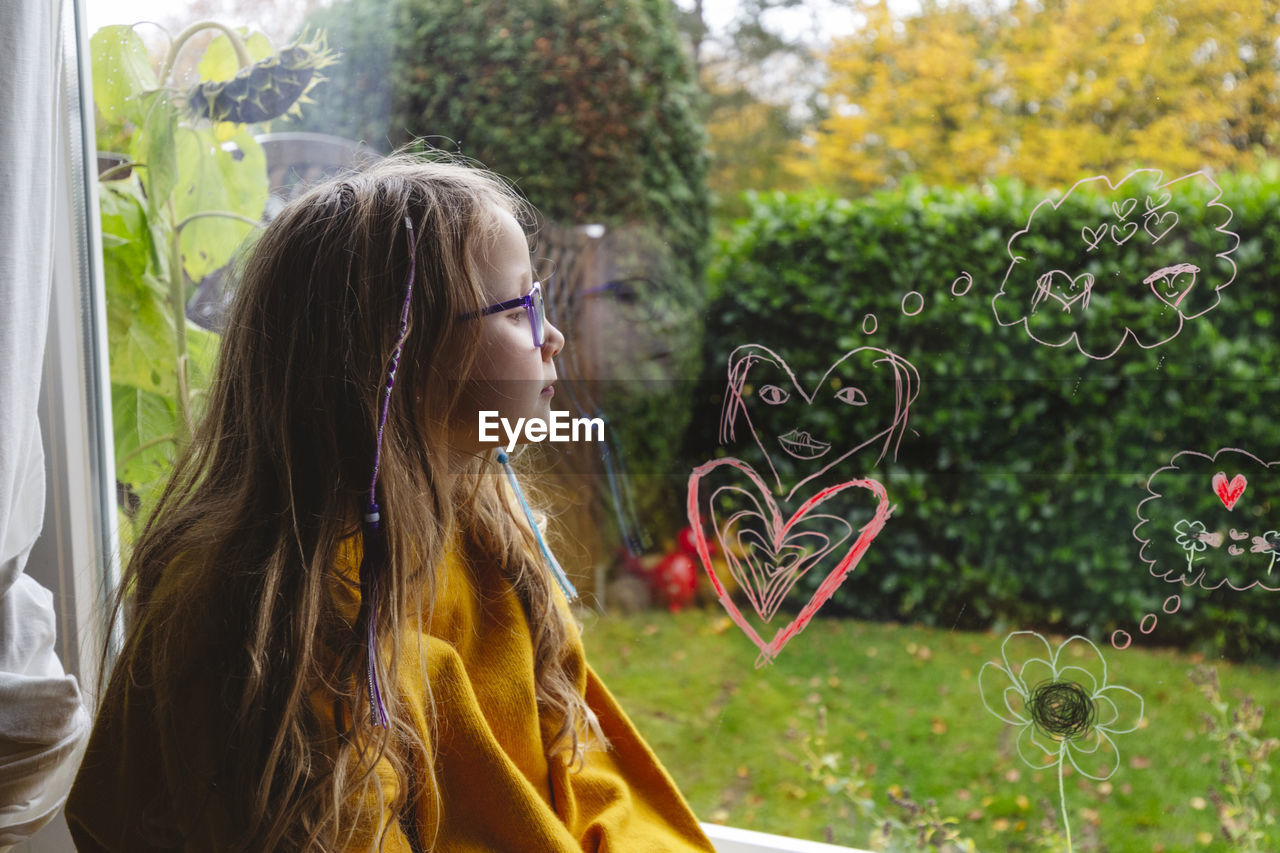 Girl in eyeglasses looking at heart shapes on transparent window at home