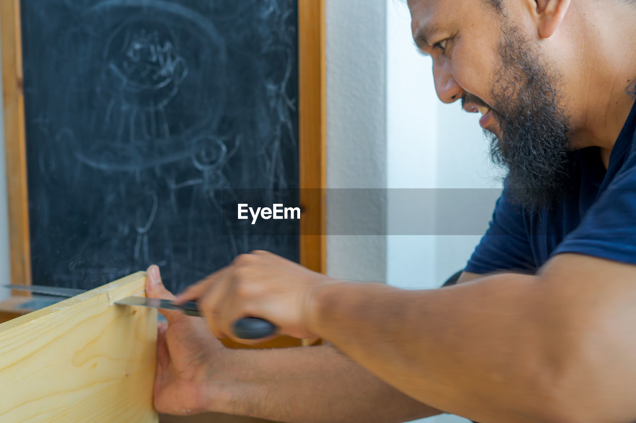 Bearded asian carpenter using hand saw cutting wooden boards