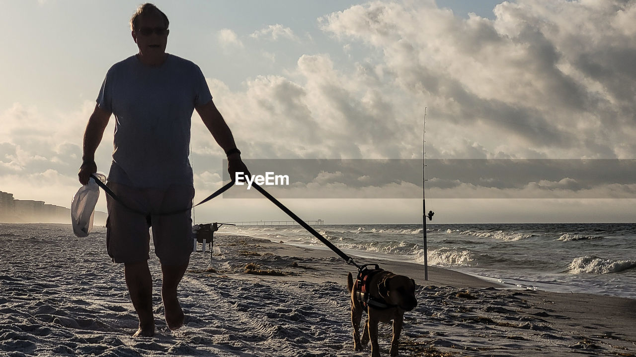 dog, sky, water, sea, beach, animal, one person, canine, land, one animal, nature, ocean, domestic animals, adult, animal themes, pet, mammal, wave, sunset, sand, person, men, full length, carnivore, coast, beauty in nature, cloud, walking, standing, motion, lifestyles, leash, shore, activity, silhouette, rear view, sports, leisure activity, clothing, pet leash, outdoors, sunlight, back lit, holding, sun, scenics - nature