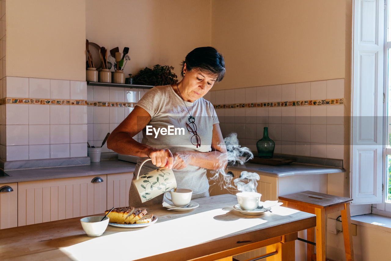 Mature female pouring hot tea from ceramic teapot into cup at table with homemade cake in cozy kitchen with wooden furniture in sunlight
