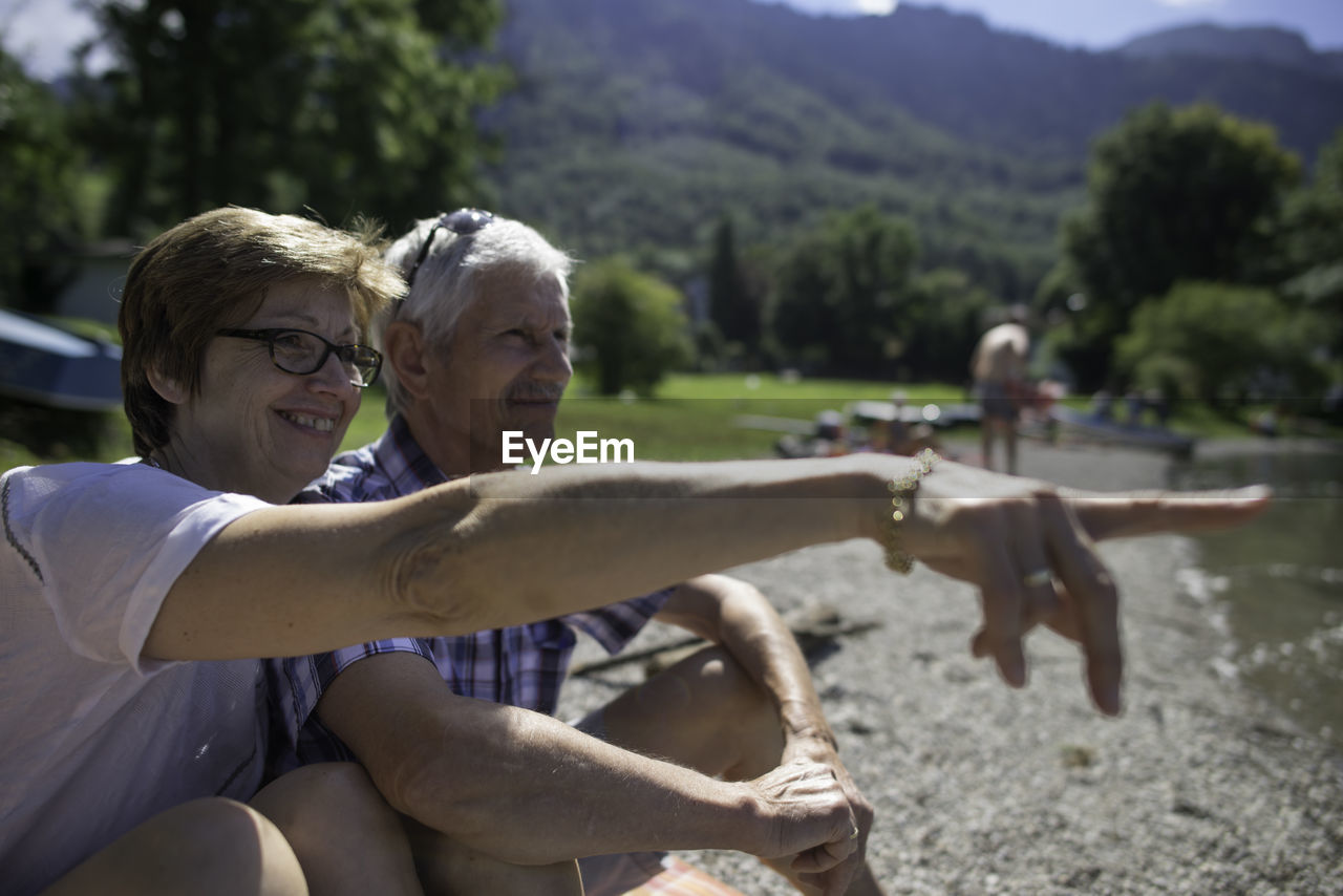 Senior woman pointing to husband while sitting at kochelsee lakeshore