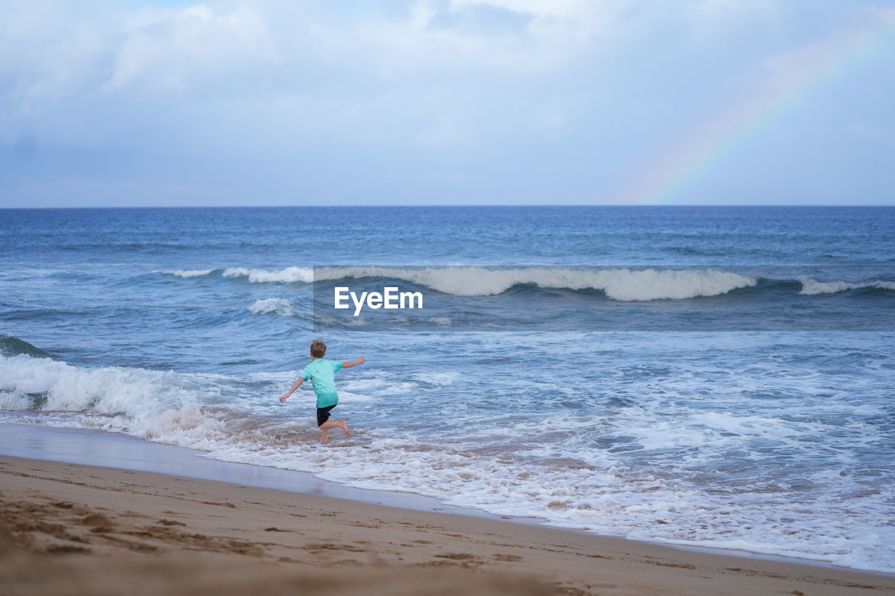 Young boy playing in the waves with a rainbow over the ocean on ka'anapali beach in hawaii. 