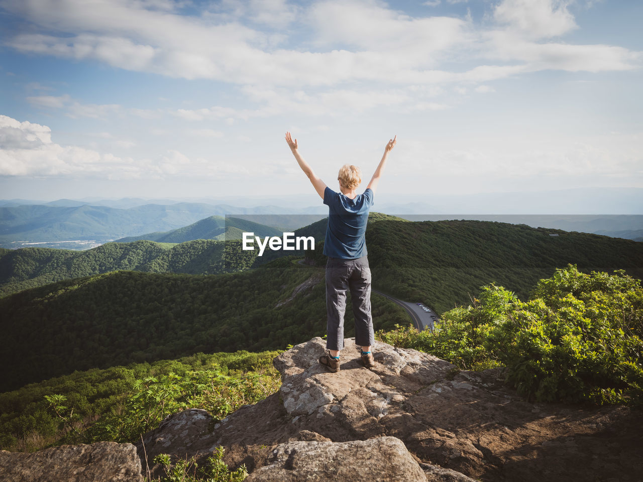 REAR VIEW OF MAN STANDING ON ROCK AGAINST MOUNTAIN