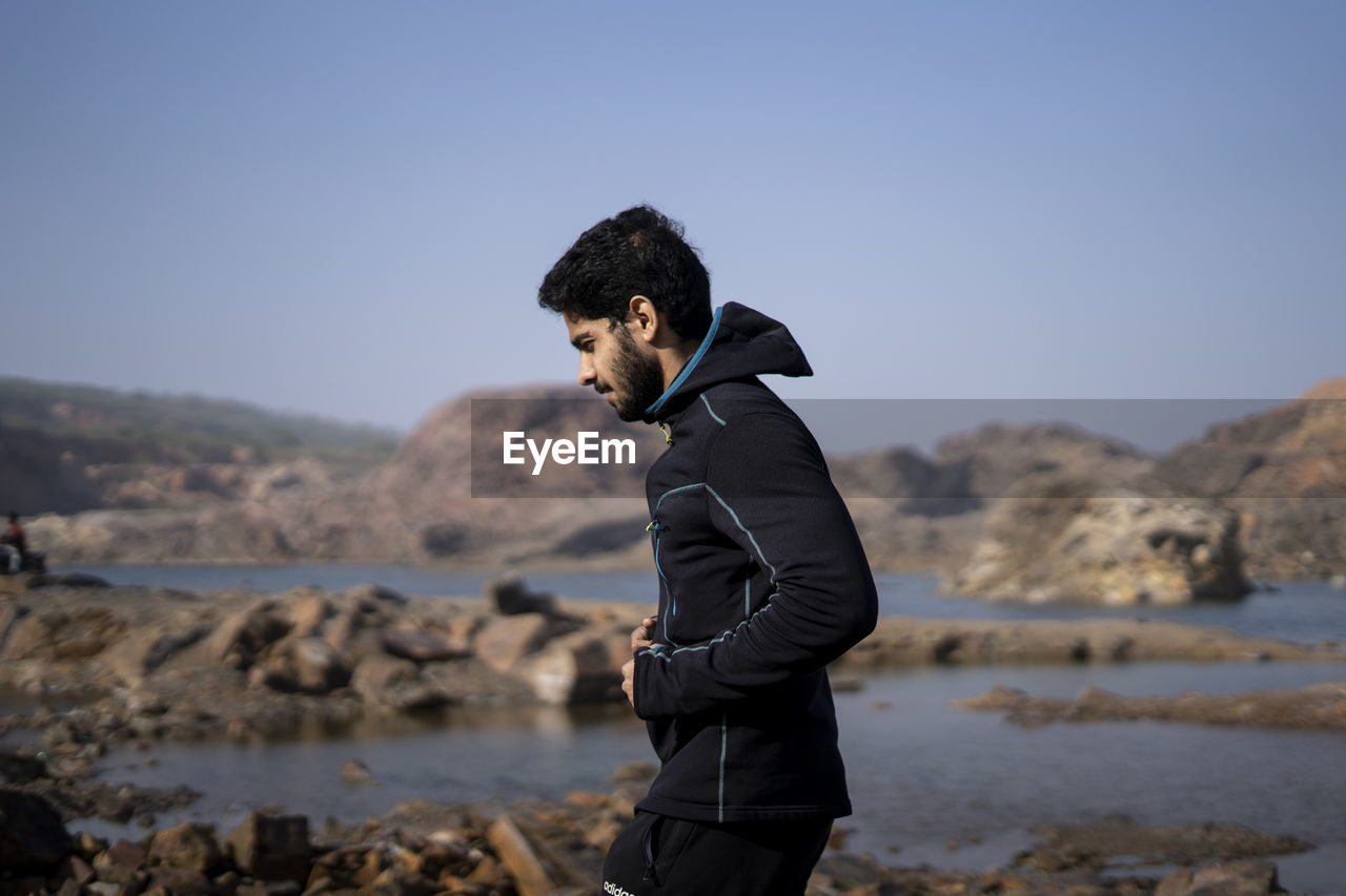 SIDE VIEW OF YOUNG MAN STANDING ON ROCK AGAINST SKY