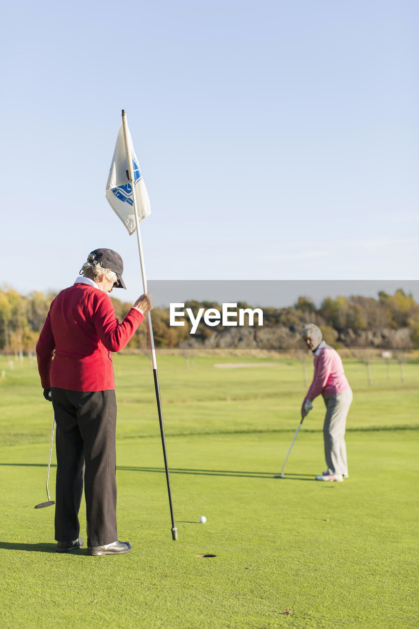 Senior woman putting while friend holding flag on golf course