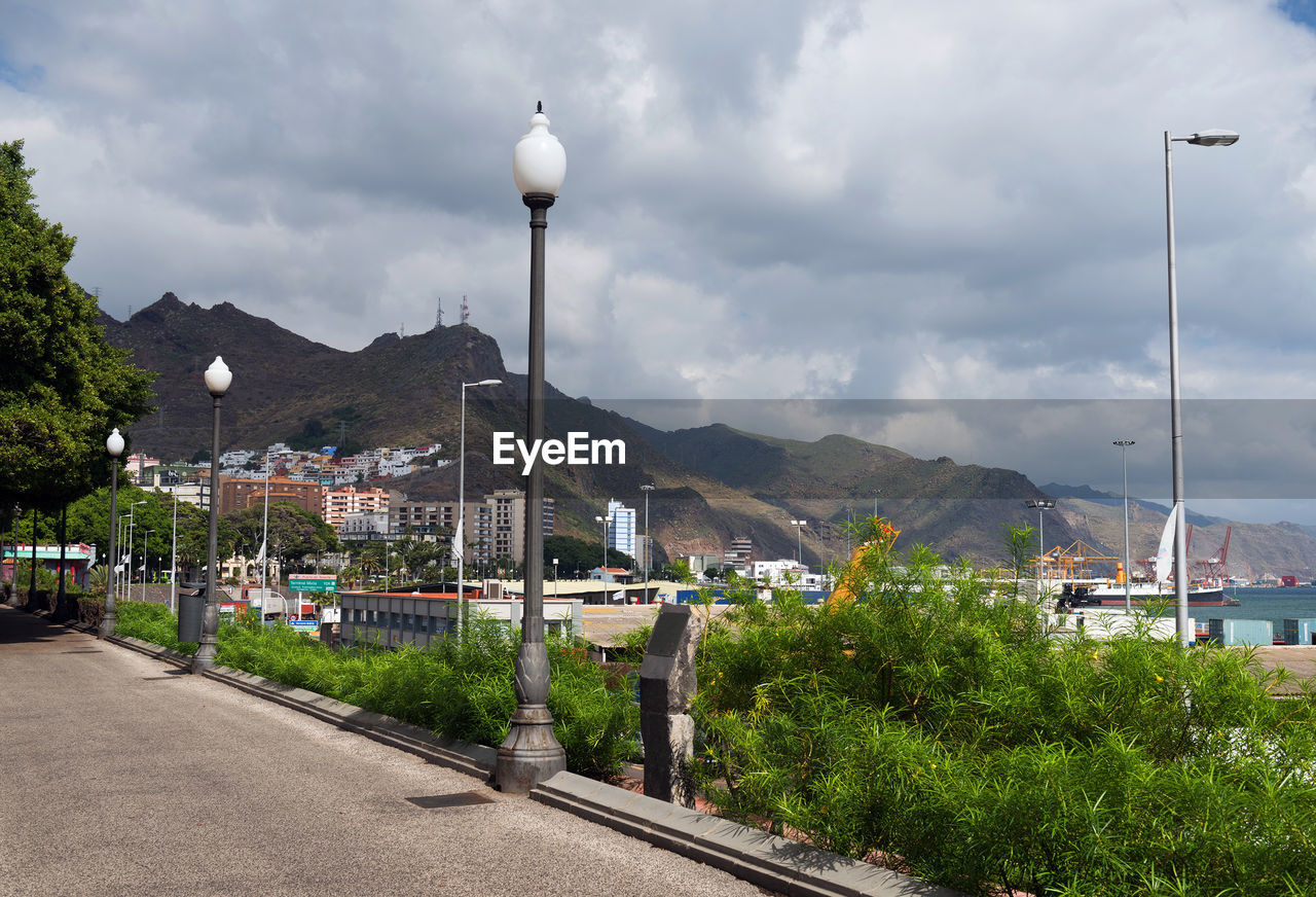 Plants and lamp posts by walkway against cloudy sky