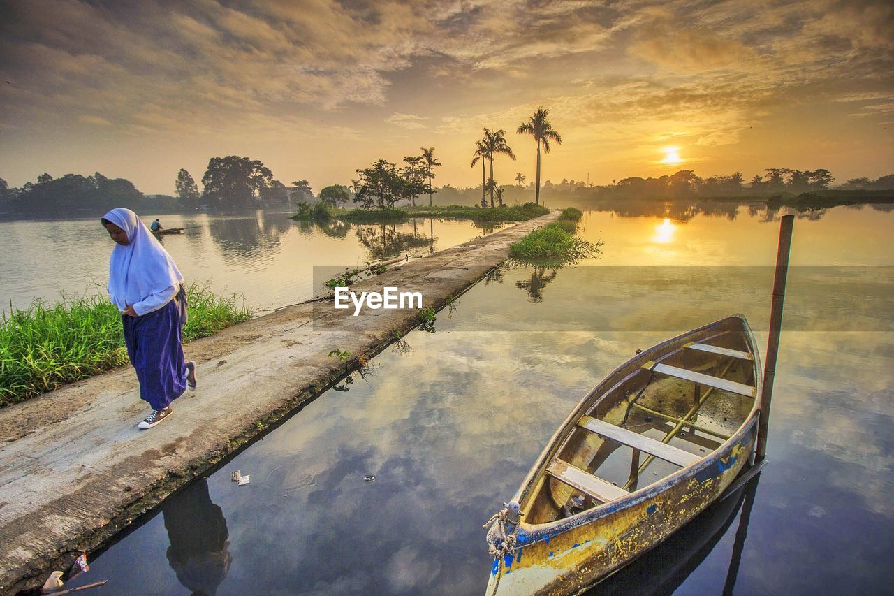 REAR VIEW OF MAN STANDING BY LAKE AGAINST SKY DURING SUNSET