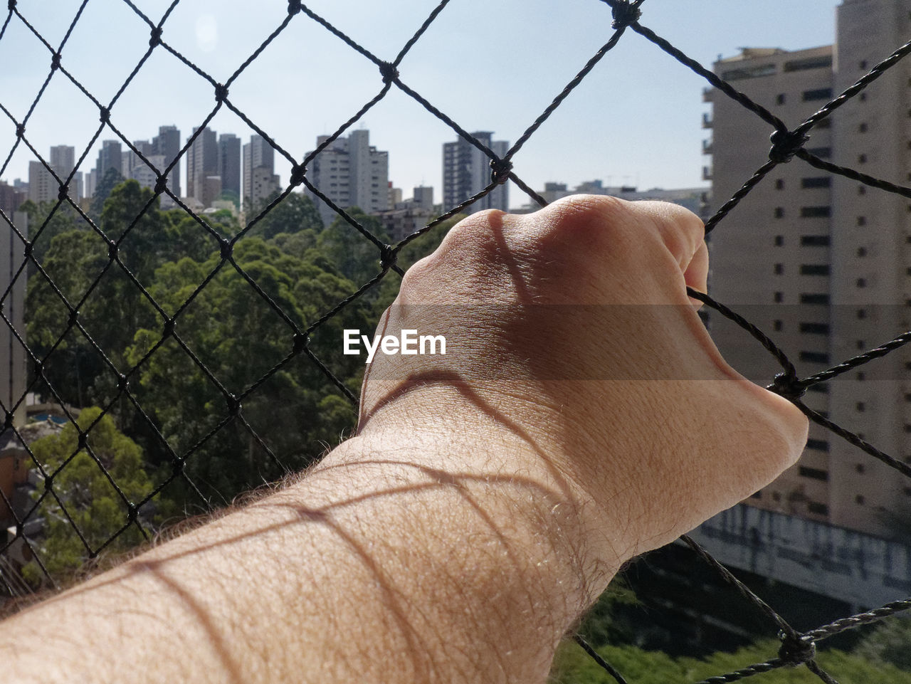 MIDSECTION OF MAN WITH CHAINLINK FENCE AGAINST CITYSCAPE SEEN THROUGH METAL