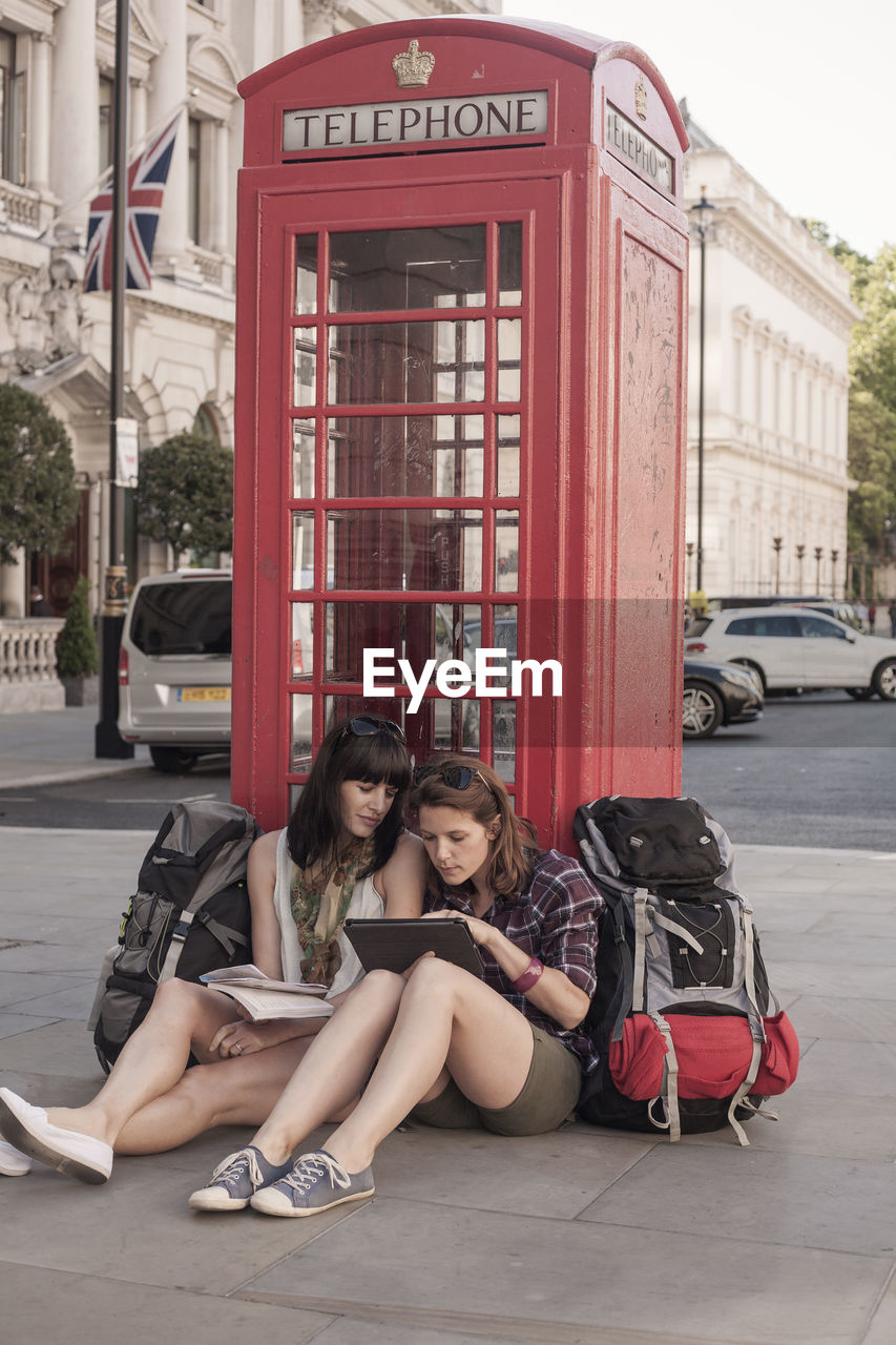 Female backpackers sitting with digital tablet against red telephone box in city