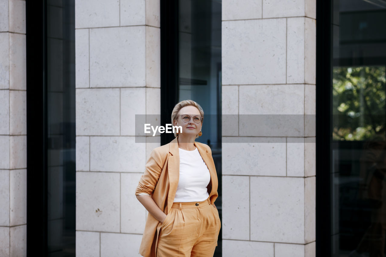 Businesswoman with hands in pockets looking away while standing against wall