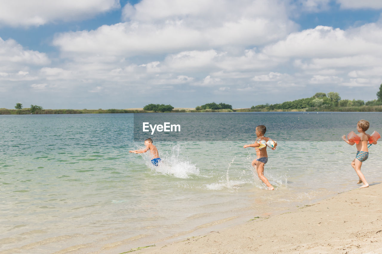 Side view of shirtless boys running towards sea against cloudy sky