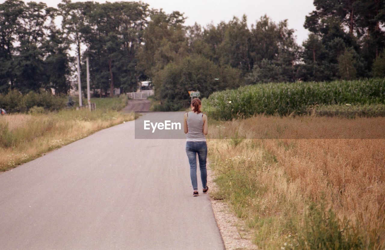 WOMAN STANDING ON ROAD AMIDST TREES AGAINST PLANTS