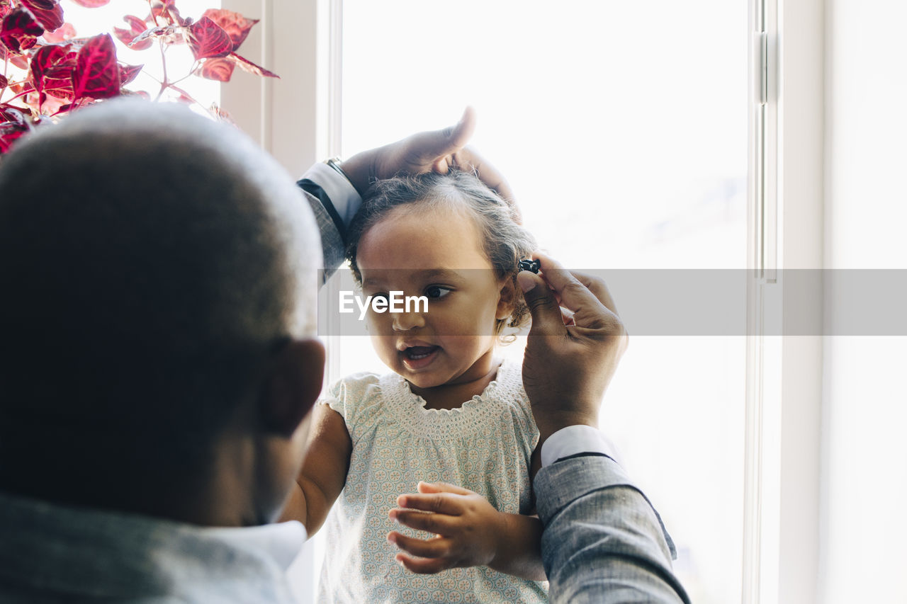 Rear view of father tying daughter's hair against window at home