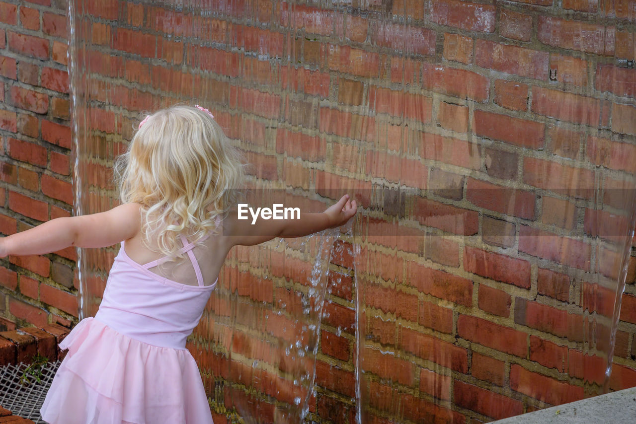 Rear view of girl touching artificial waterfall over brick wall