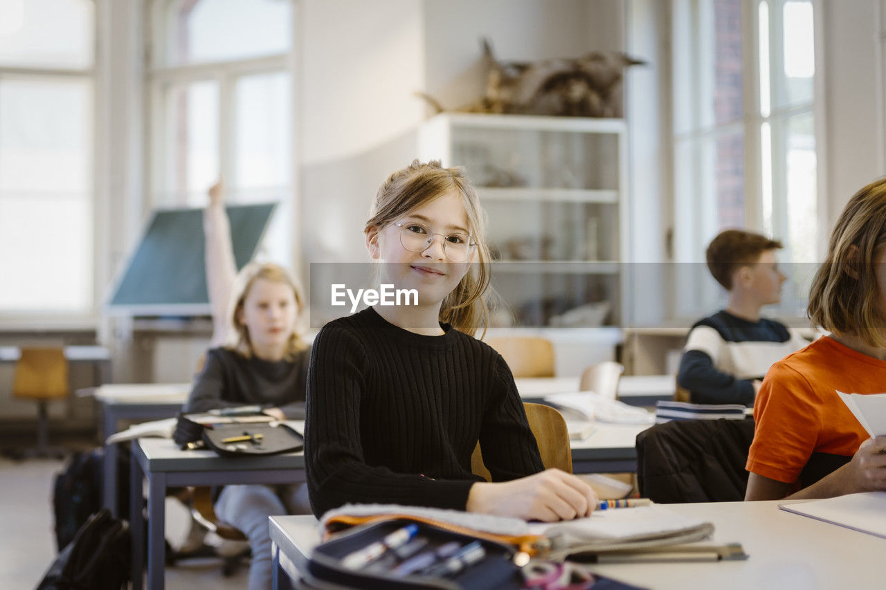 Portrait of smiling schoolgirl sitting at desk with friend in classroom