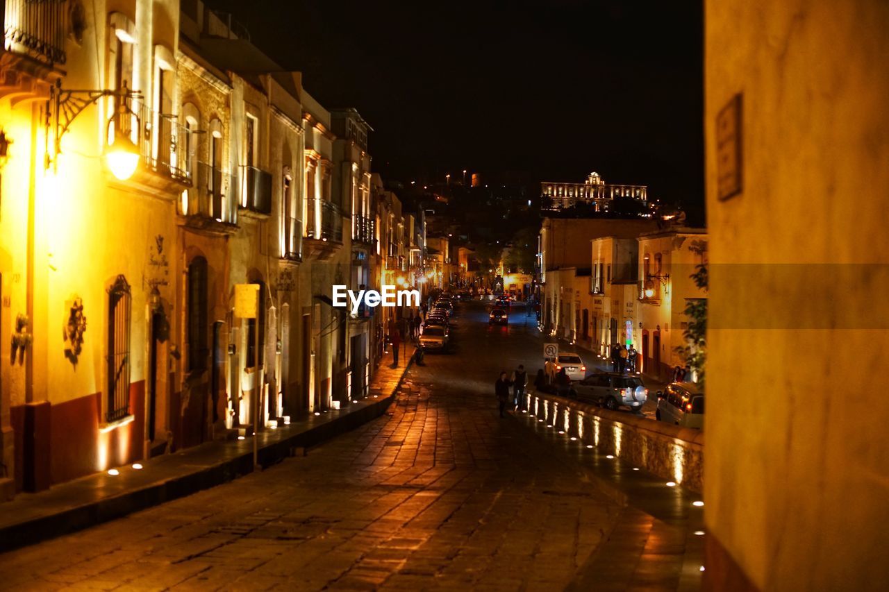 ILLUMINATED STREET BY BUILDINGS IN CITY AT NIGHT