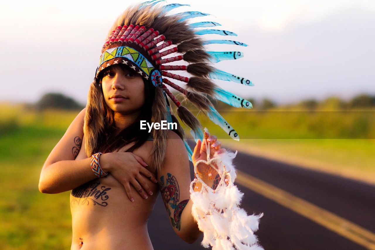 Portrait of shirtless woman in headdress standing against sky