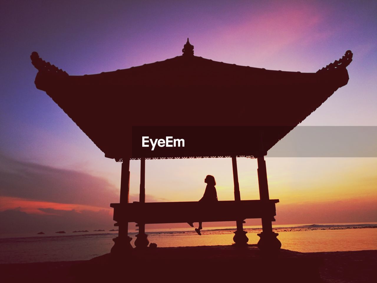 Silhouette woman sitting on bench at beach against sky during sunset