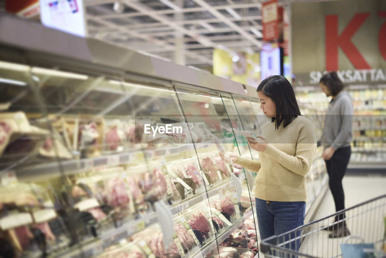 Woman doing shopping in supermarket and using cell phone to compare prices or checking shopping list