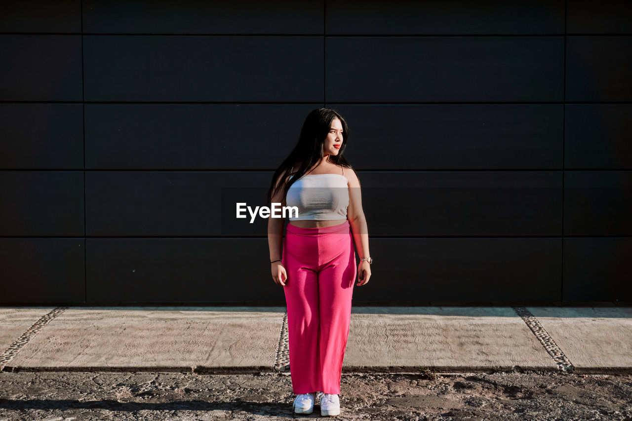 Full body chubby young woman in stylish pink pants and white top looking away while standing against black wall of contemporary building on sunlit city street