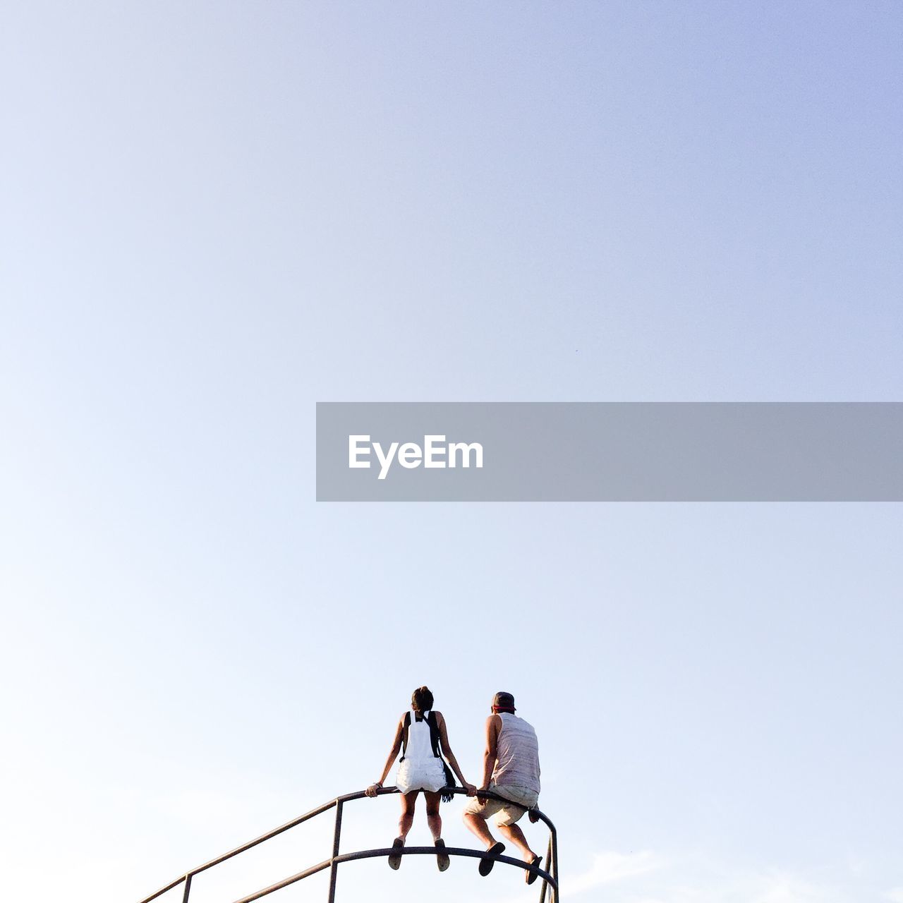 Low angle view of man and woman sitting against clear sky on sunny day