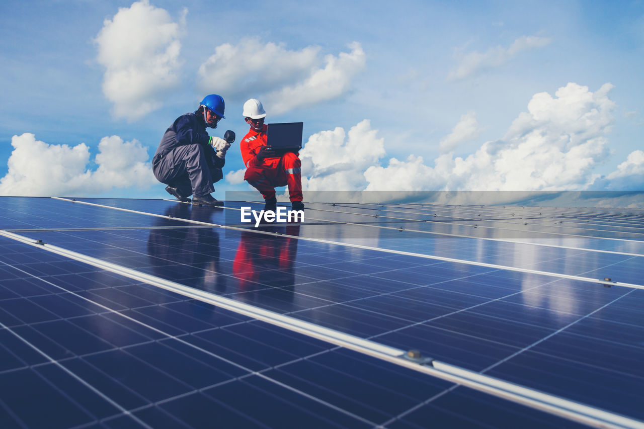 Low angle view of male technicians working on solar panels against sky