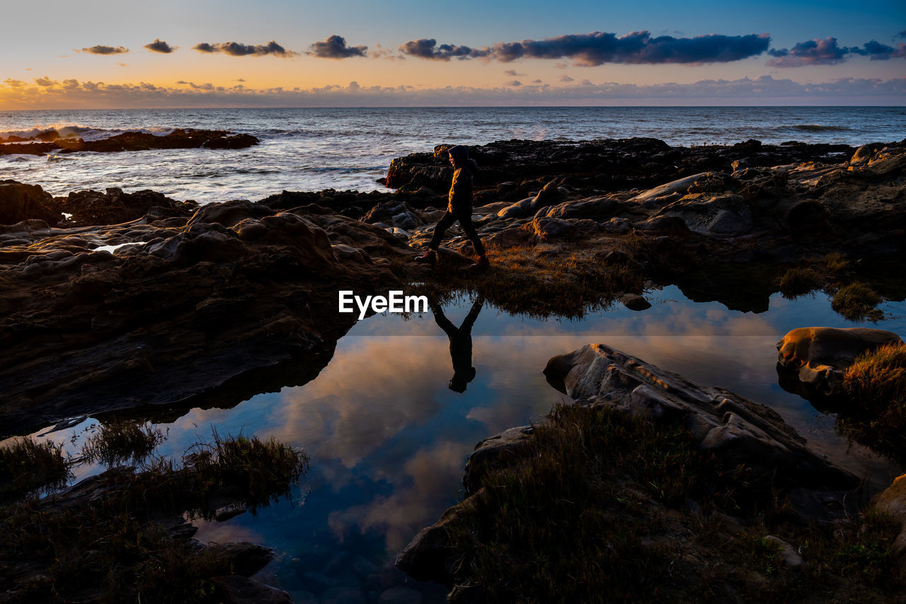 Teen walking on rocky shore casting reflection under sunset sky