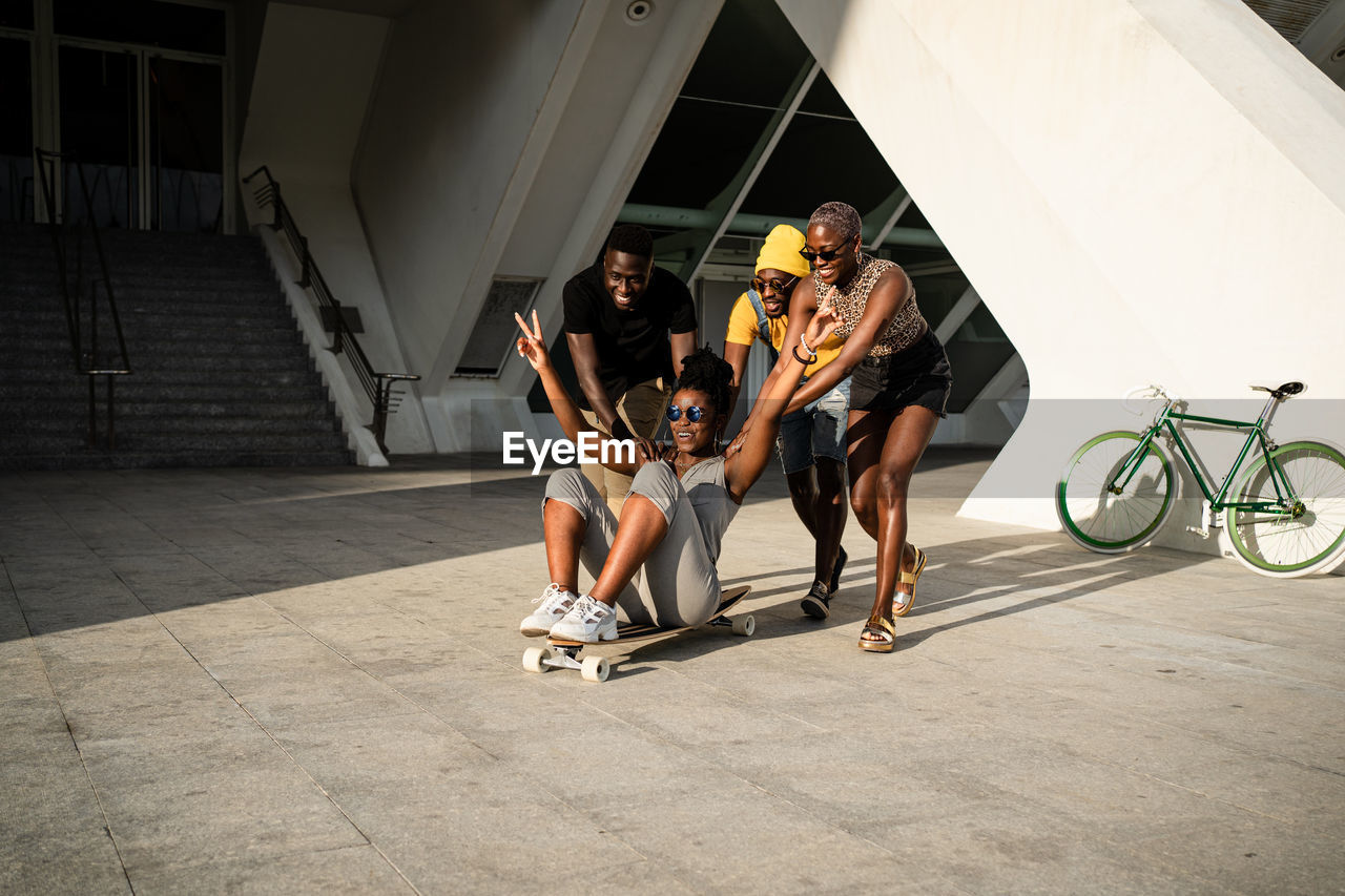 Company of positive african american friends riding woman on longboard while having fun together on street in summer