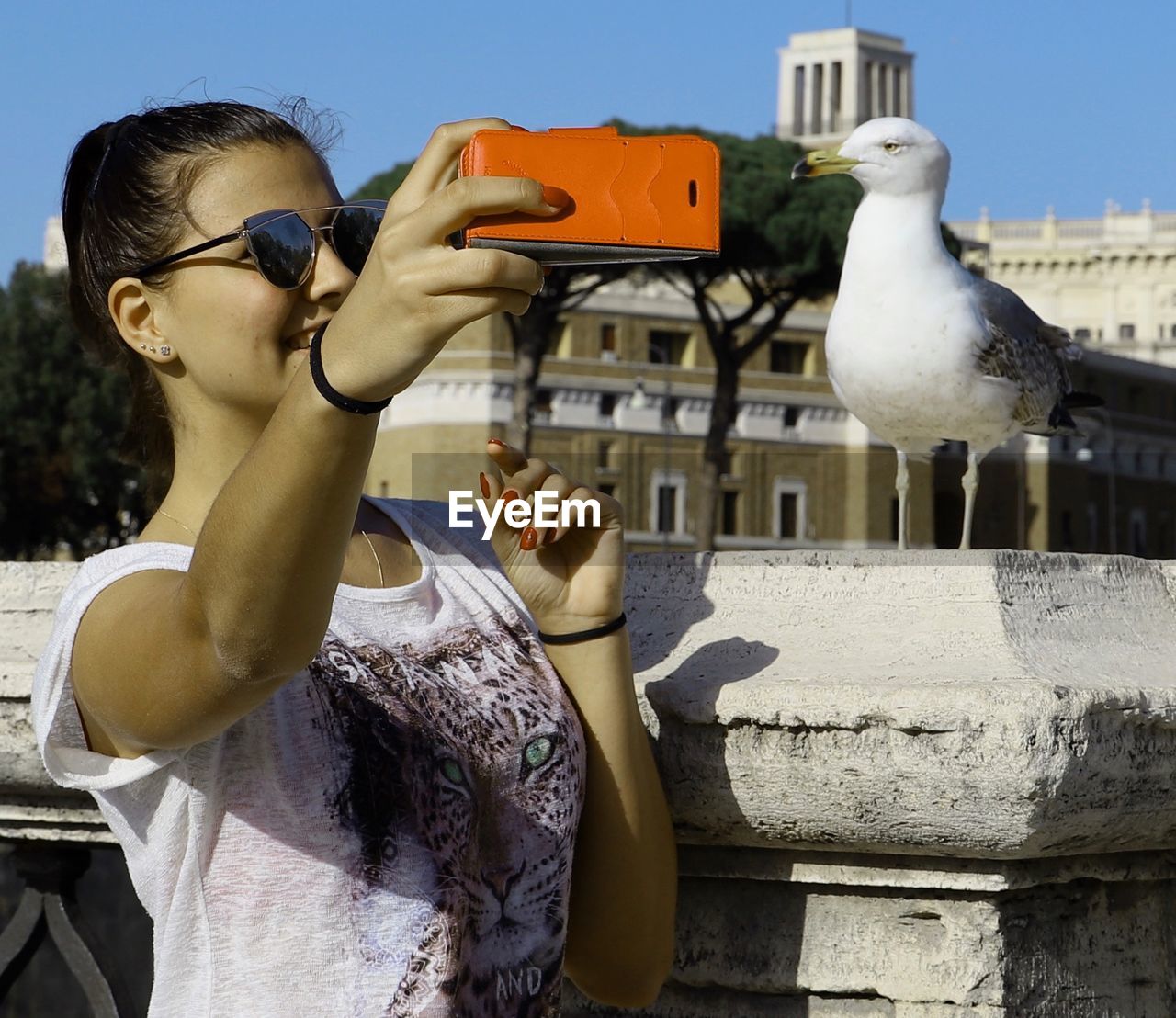 Happy young woman taking selfie through smart phone with seagull perching on railing