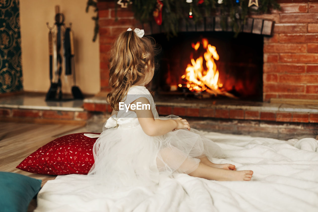 A happy little girl is sitting by the fireplace in a cozy living room on christmas eve