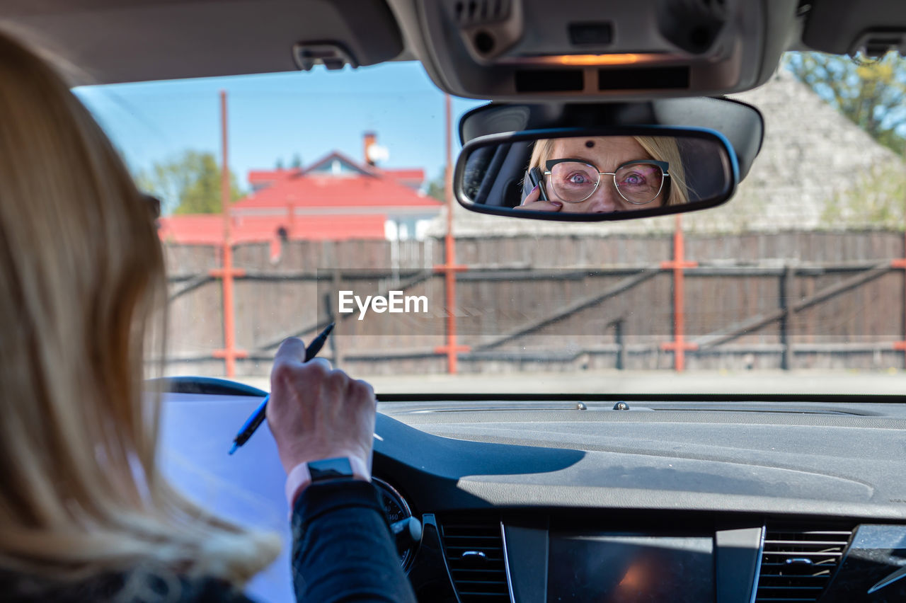 Middle-aged woman in glasses in the car working with documents and talking on the phone, rear view