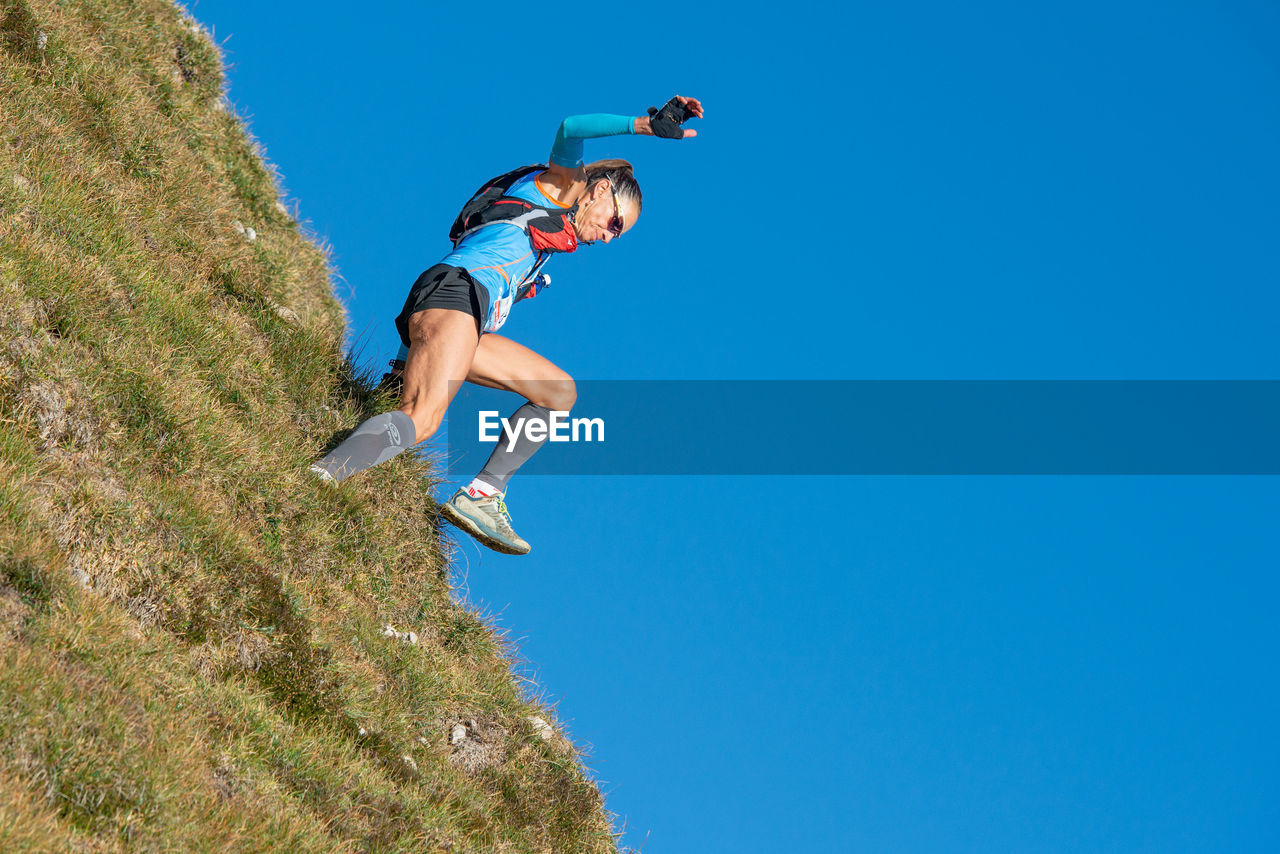 Low angle view of man jumping on mountain against clear blue sky