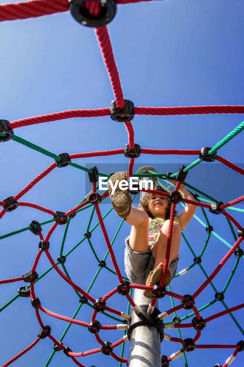 Low angle view of cute girl climbing on rope pyramid against clear blue sky